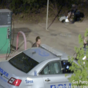 The male suspect stands behind a police vehicle while an officer searches his belongings in the background. The male suspect stands behind a police vehicle while an officer searches his belongings in the background.